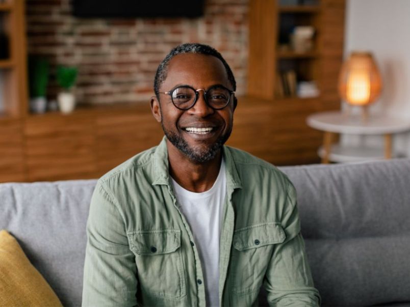 A middle-aged man in glasses and a green button-up sitting on a couch smiling